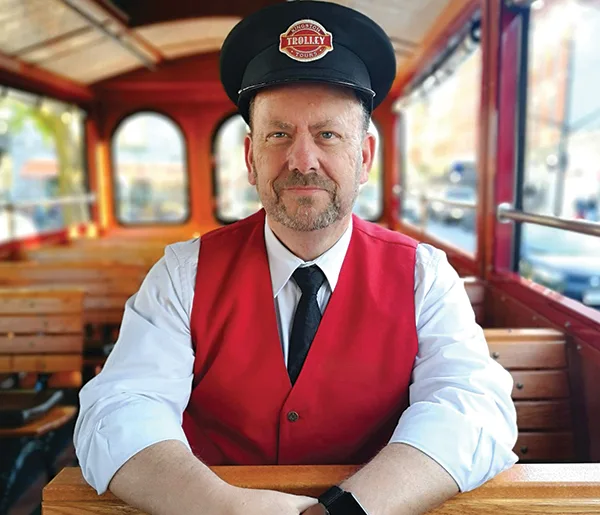 Trolley Driver in a red vest and black driving cap sitting on the wooden benches inside the classic red Kingston Trolley.